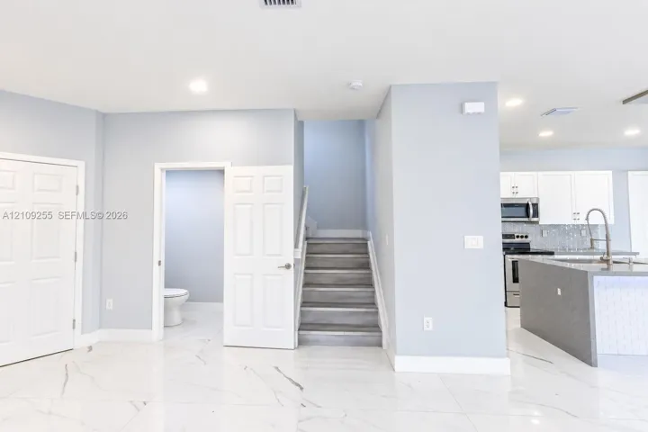 Large-format white tile flooring with grey veining, staircase with wood-finish steps, and a kitchen featuring white cabinetry, a center island with a stone-finish countertop, and stainless steel appliances
