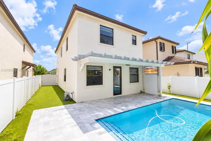 Stucco exterior featuring a pergola-covered patio, paved pool deck, private swimming pool, and artificial turf lawn enclosed by white vinyl fencing
