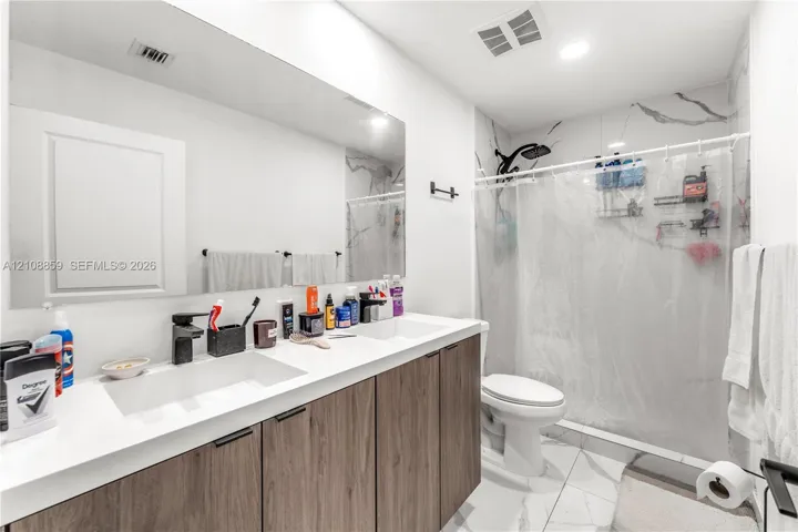 Double vanity featuring wood-finish cabinetry, rectangular under-mount sinks, matte black faucets, and an expansive wall-to-wall mirror, complemented by tiled flooring and a tiled shower surround