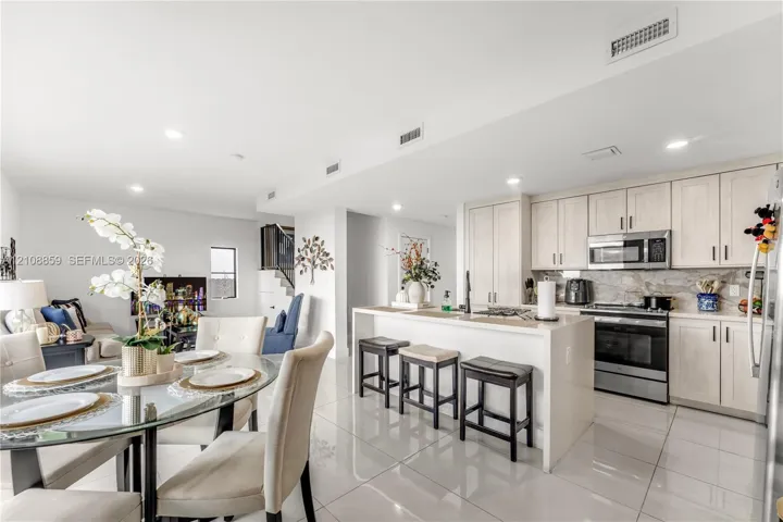 Open-concept kitchen featuring light-toned cabinetry, stainless steel appliances, a stone-finish backsplash, a central island with integrated seating, and high-gloss tile flooring