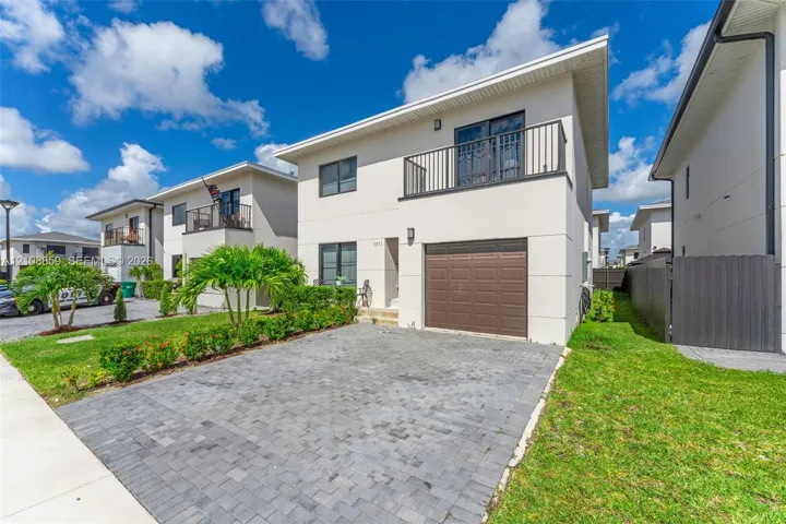 Two-story exterior featuring a smooth stucco finish, a private second-level balcony with metal railings, a stone-paver driveway, and an attached single-car garage