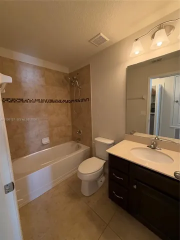 Bathroom featuring a dark wood vanity with an integrated basin, a neutral-toned countertop, a shower-tub combination with tile surround and decorative inlay, and wall-mounted vanity lighting