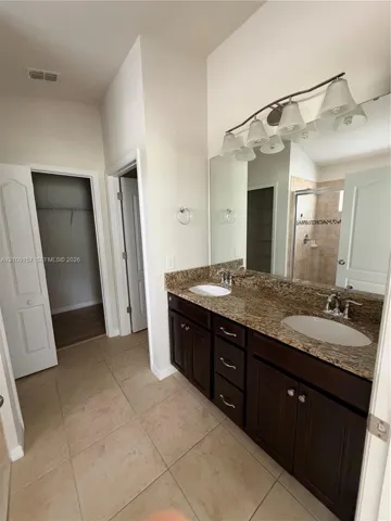 Dual-sink vanity with stone countertop, dark wood-finish cabinetry, full-width vanity mirror, polished metal lighting fixture, and tile flooring