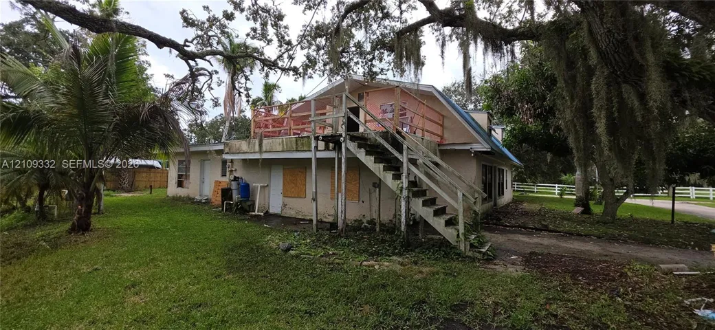 Two-story residential structure featuring an elevated wood deck, exterior staircase, and mature tree canopy on the grounds