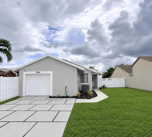 Single-story residence featuring a paved grid driveway, stucco exterior, attached garage, manicured lawn, and white vinyl privacy fencing