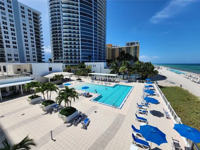 Expansive paved pool deck featuring a swimming pool, integrated landscaping with palms, and direct beachfront access