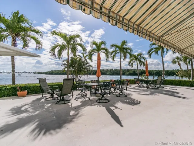 Spacious outdoor patio with water frontage, bordered by a manicured hedge and mature palm trees