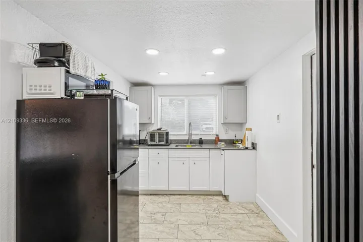 Kitchen featuring white shaker-style cabinetry, solid-surface countertops, stone-finish tile flooring, recessed ceiling lighting, and a stainless steel kitchen sink