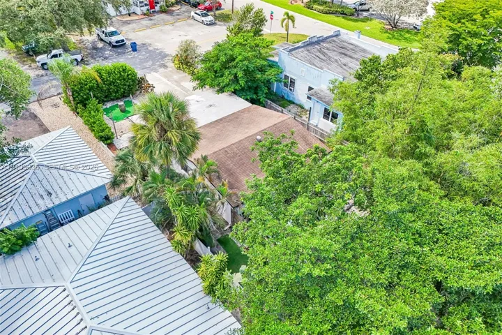 Aerial view of the property featuring a metal roofing system, shingle-roof structure, mature trees, palm landscaping, and synthetic grass ground cover