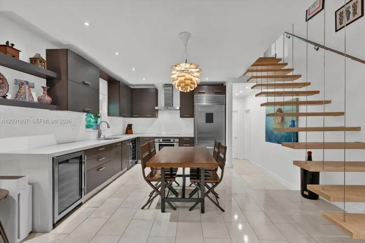 Custom cabinetry with horizontal hardware, light-toned stone tile flooring, stainless steel integrated refrigeration, stone-surfaced countertops, and an open-tread staircase with steel cable railing
