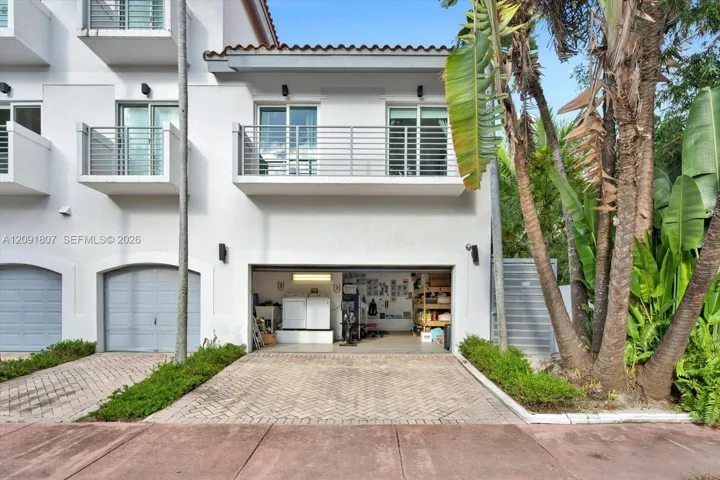 Stucco exterior featuring dual-level metal balconies, terracotta-style roof tile accents, a private herringbone-patterned paver driveway, and an attached multi-car garage