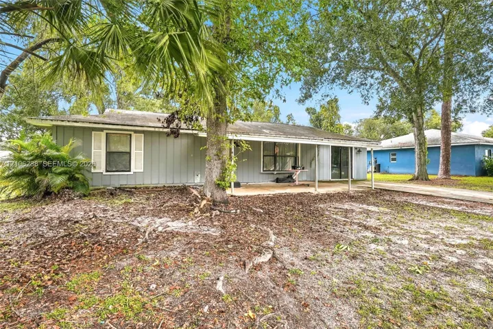 Single-story residence featuring vertical siding, a covered concrete patio, and a shingled roof, set on a lot with mature trees and palms