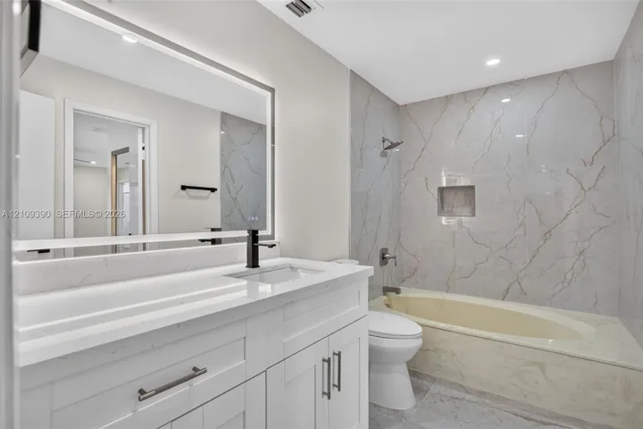Bathroom featuring a vanity with a white countertop, integrated rectangular sink, matte black faucet, and a large LED-backlit mirror, complemented by a tub enclosure with full-height stone-finish wall tile and a built-in recessed niche
