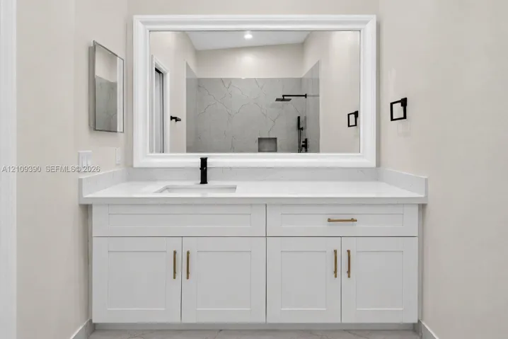 White shaker-style cabinetry with brass hardware, solid surface countertop, rectangular undermount sink, matte black faucet, and large framed wall mirror
