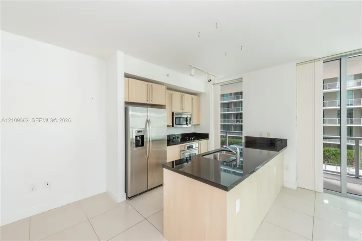 Kitchen featuring light wood-finish cabinetry, dark solid-surface countertops, stainless steel refrigerator, built-in microwave, and large-format tile flooring