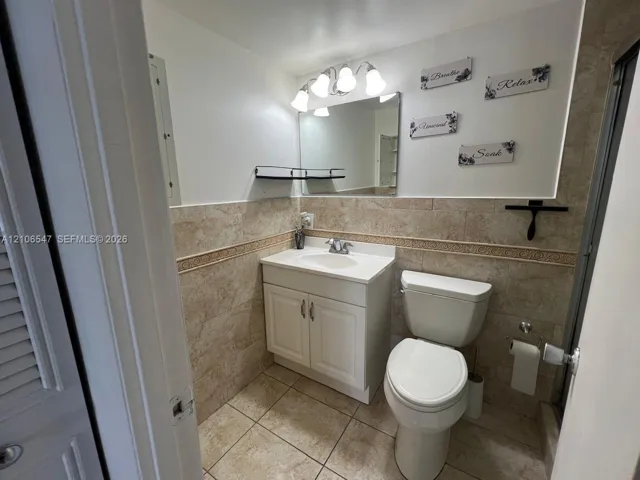 Bathroom featuring stone-finish wall and floor tile with decorative border, white cabinet vanity with integrated sink, four-light vanity fixture, and rectangular wall mirror