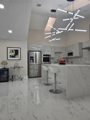 Modern kitchen featuring a waterfall island with stone-finish countertops, high-gloss cabinetry, stainless steel appliances, large-format tile flooring, and geometric multi-pendant lighting