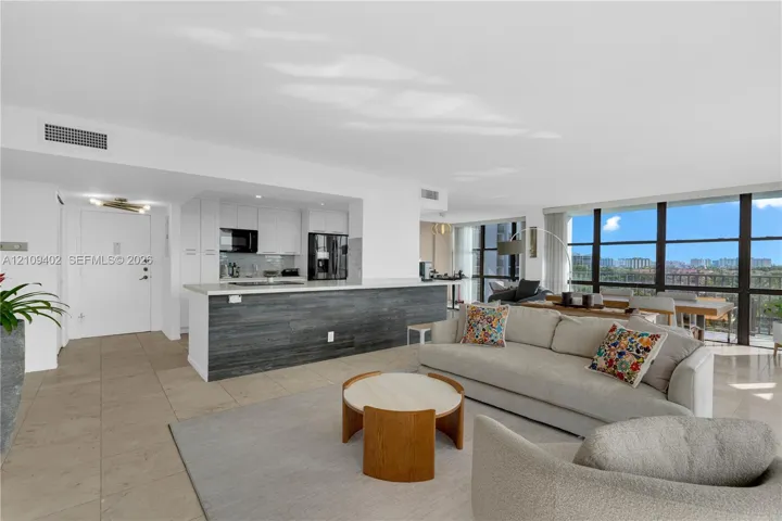 Open-concept interior featuring large-format tile flooring, a kitchen island with stone-finish countertop, dark wood-grain cabinetry accents, and floor-to-ceiling windows