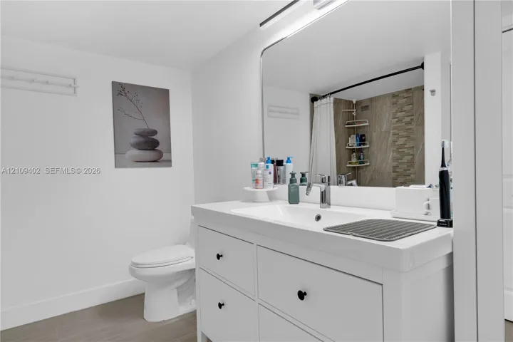 Bathroom featuring a white vanity cabinet with a rectangular basin, large wall-mounted mirror, wood-finish flooring, and a tiled shower surround with decorative stone-finish accents