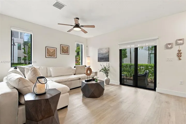 Neutral-toned living space featuring light-colored wood-finish flooring, a ceiling fan with integrated lighting, dual vertical windows, and a sliding glass door providing external access