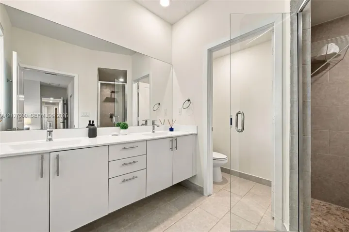 Bathroom featuring a dual-sink floating vanity with white countertops, expansive wall-to-wall mirror, glass-enclosed shower, and neutral tile flooring