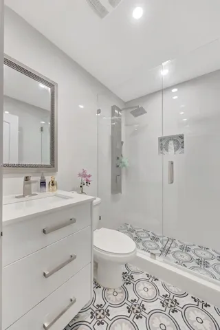 White vanity with a rectangular sink, brushed nickel hardware, patterned tile flooring, walk-in shower with a glass enclosure, and a wall-mounted shower panel