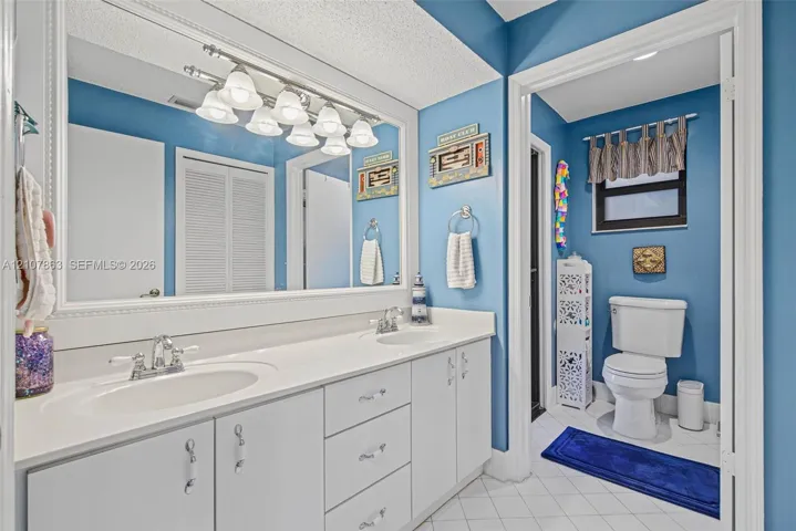 Dual-sink vanity featuring a solid surface countertop, white cabinetry with polished hardware, a large wall-mounted mirror, and a multi-bulb light fixture
