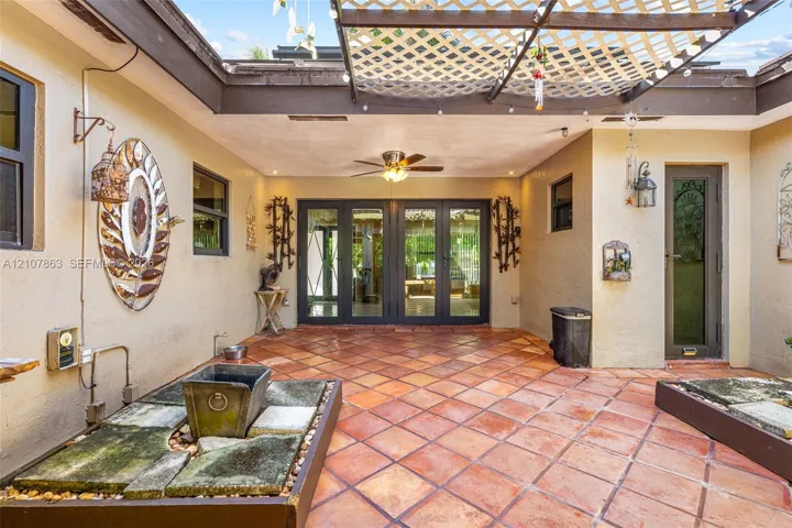 Enclosed courtyard featuring terracotta tile flooring, a structured wooden lattice overhead, multiple built-in stone planters, and a ceiling-mounted fan