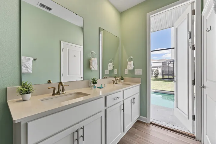 Dual-sink vanity with light-toned countertop, white cabinetry with metal hardware, wall-to-wall mirrors, and wood-finish flooring, featuring a door providing exterior access to a pool and screened patio area