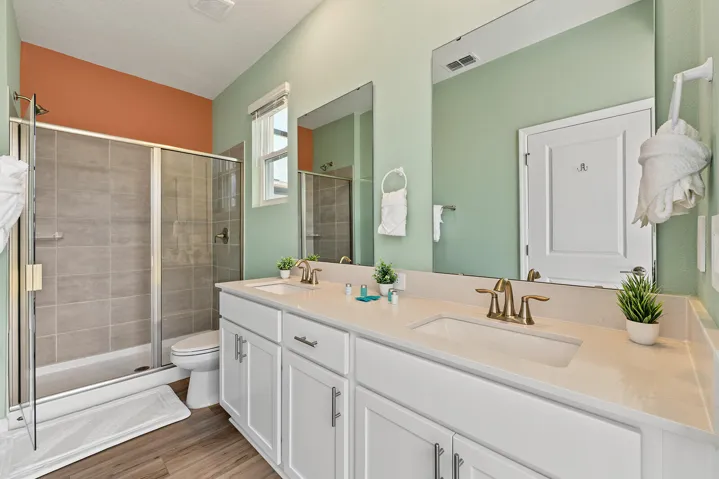 Double vanity with quartz countertops, undermount sinks, bronze-finish fixtures, and full-height mirrors, accompanied by a glass-enclosed walk-in shower with tile surround and wood-finish flooring