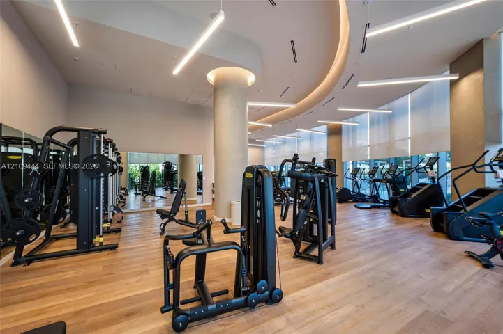 Fitness space featuring wood-finish flooring, floor-to-ceiling windows with roller shades, recessed cove lighting, suspended linear light fixtures, and a structural column