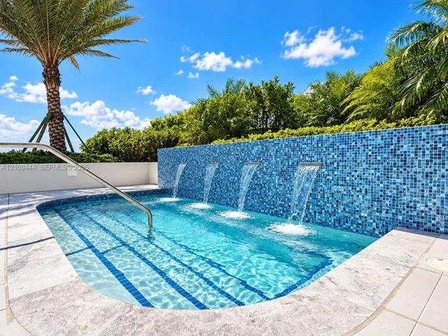 In-ground pool featuring a blue glass mosaic tile water wall, four integrated water features, a metal handrail, and stone-finish coping