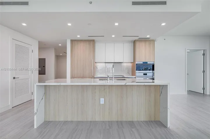 Kitchen featuring a center island with a stone-finish waterfall countertop, custom wood-finish cabinetry, white matte upper cabinets, integrated stainless steel appliances, and neutral flooring