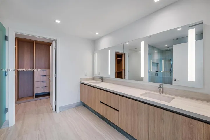 Floating double vanity with wood-finish cabinetry, integrated rectangular sinks, expansive wall-to-wall mirror with built-in vertical lighting, and large-format tile flooring
