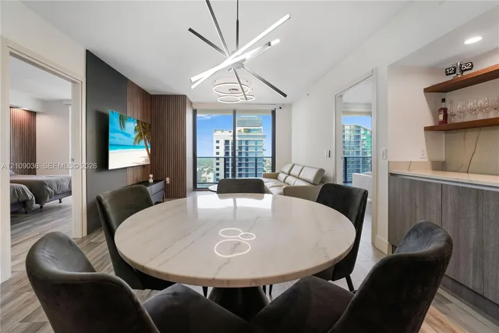 Modern living space featuring floor-to-ceiling windows, wood-panel accent wall, built-in wet bar with shelving, integrated recessed lighting, and a linear architectural chandelier