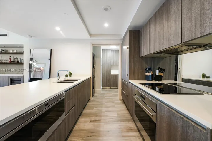 Modern kitchen featuring wood-finish cabinetry, white countertops, built-in stainless steel appliances, a mirrored backsplash, and wood-finish flooring