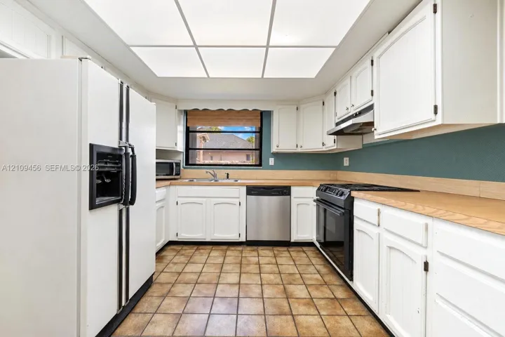 Kitchen featuring white cabinetry, light-toned countertops, ceramic tile flooring, a stainless steel dishwasher, and a recessed tray ceiling with integrated lighting