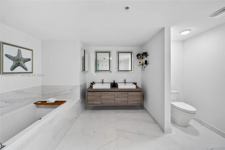 Floating wood-finish vanity with dual vessel sinks, large-format stone-tile flooring, integrated soaking tub, and wall-mounted mirrors with integrated lighting
