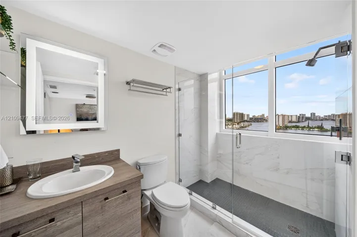 Bathroom featuring a wood-finish vanity with a drop-in sink, illuminated wall mirror, glass-enclosed walk-in shower with stone-style wall tile, and dark mosaic floor tile