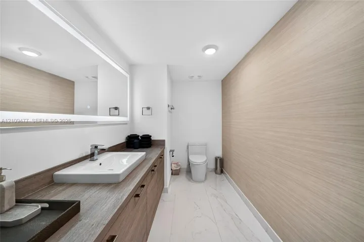 Floating vanity with wood-finish cabinetry, rectangular vessel sink, expansive integrated mirror with perimeter lighting, and light-toned tile flooring