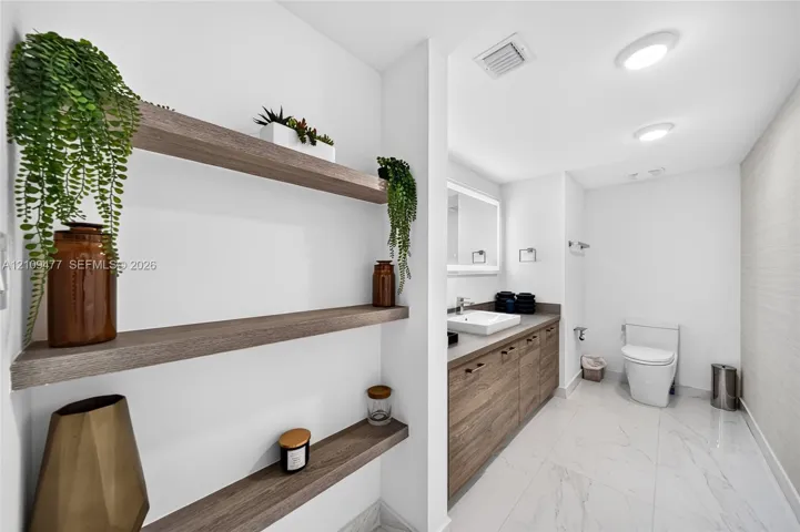 Bathroom featuring wood-finish floating shelves, wood-finish cabinetry, a square vessel sink, a backlit mirror, and stone-finish tile flooring
