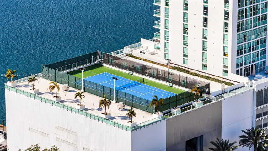 Rooftop tennis court featuring a hard surface, perimeter fencing, overhead lighting, and containerized landscaping