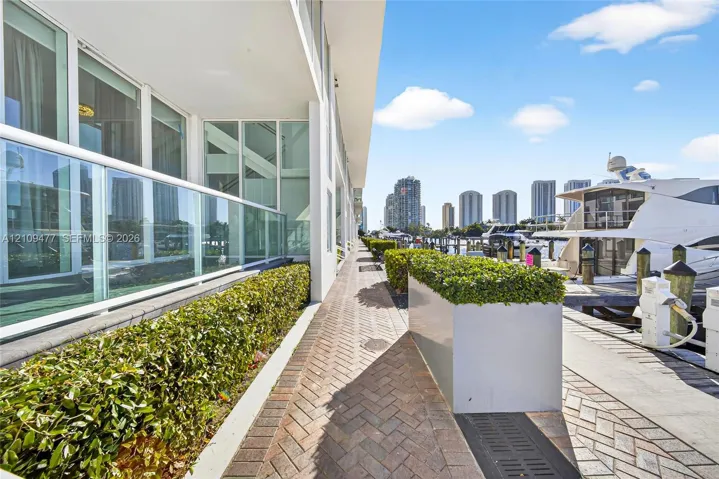Waterfront terrace featuring a herringbone-patterned brick walkway, glass railings, and built-in metal planters with mature hedges