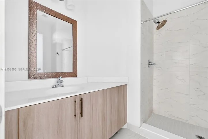 Bathroom vanity featuring a wood-finish cabinet, white solid-surface countertop, and rectangular mirror with a mosaic frame, alongside a walk-in shower with light-toned wall tile and a silver-finish shower head