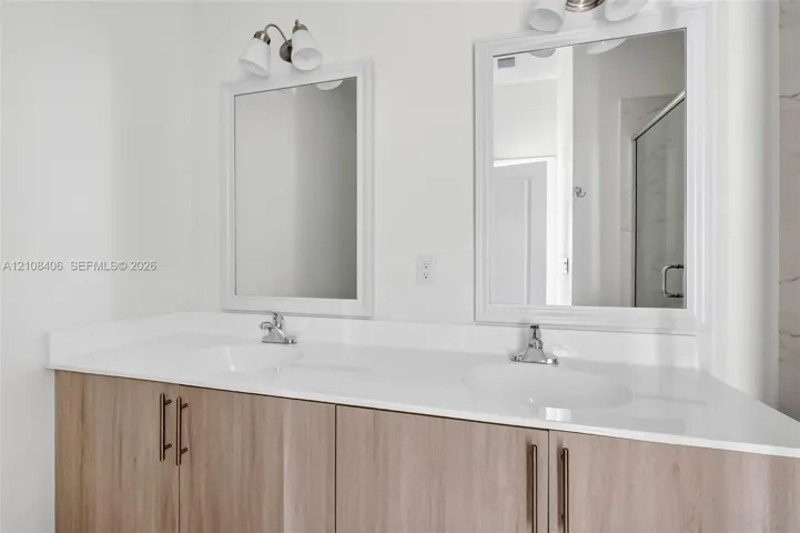 Dual-sink vanity with a white solid-surface countertop, light wood-finish cabinetry, brushed metal hardware, and framed mirrors with overhead light​​‌​​​​‌​​‌‌​‌‌​​​‌‌​‌​‌​‌​​​‌​​ fixtures