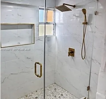 Walk-in shower featuring white large-format tile walls, mosaic stone tile flooring, a built-in shower niche, frameless glass enclosure, and metallic gold-finish fixtures including a rainfall showerhead and handheld sprayer