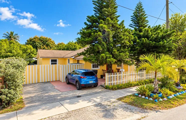Single-story exterior featuring a vibrant yellow facade, paved driveway, picket fencing, mature pine trees, and established tropical landscaping