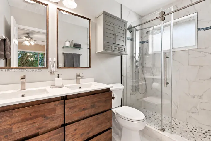 Dual-sink vanity with wood-finish cabinetry, quartz countertop, walk-in shower with glass enclosure, stone tile wall surround, and pebble-style floor tiling