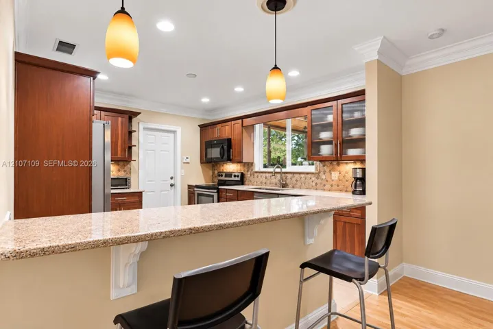 Kitchen featuring warm-tone wood cabinetry, stone-surfaced peninsula countertop, crown molding, recessed lighting, and pendant fixtures