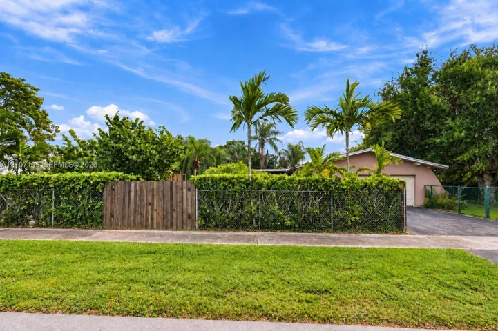 Exterior grounds featuring a metal chain-link perimeter fence, a solid wood privacy gate, mature tropical landscaping, and an asphalt driveway